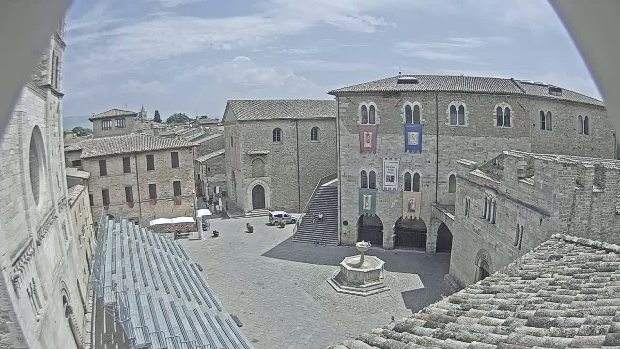 A town square with stone buildings, a fountain, and empty bleachers under a partly cloudy sky.
