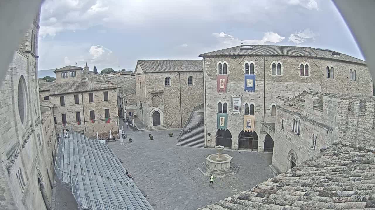 A partly cloudy day reveals a town square surrounded by medieval-style stone buildings, including a large building with banners hanging from its facade and a fountain in the center of the square.