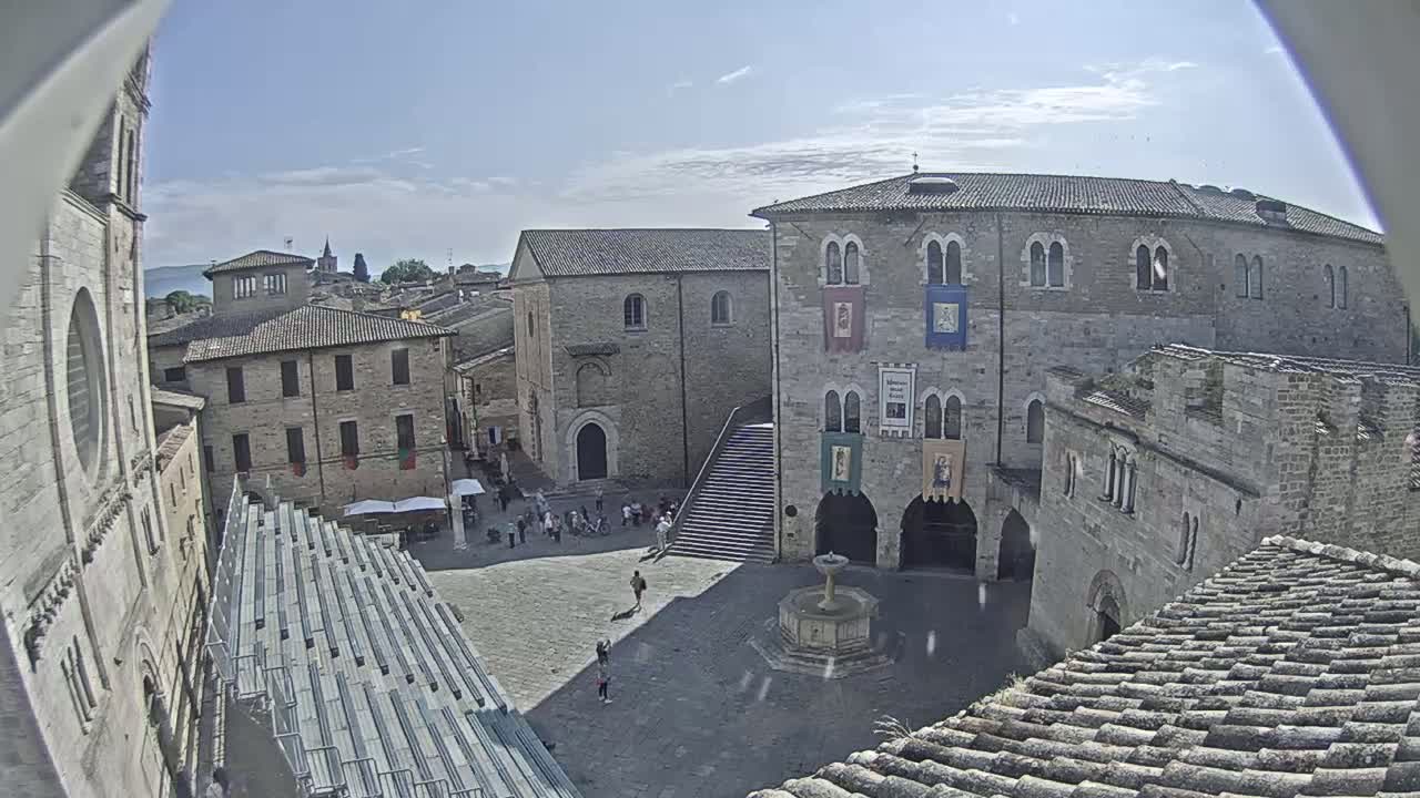 A sunny view of a town square surrounded by medieval-style buildings, a fountain in the center, and bleachers set up along one side.