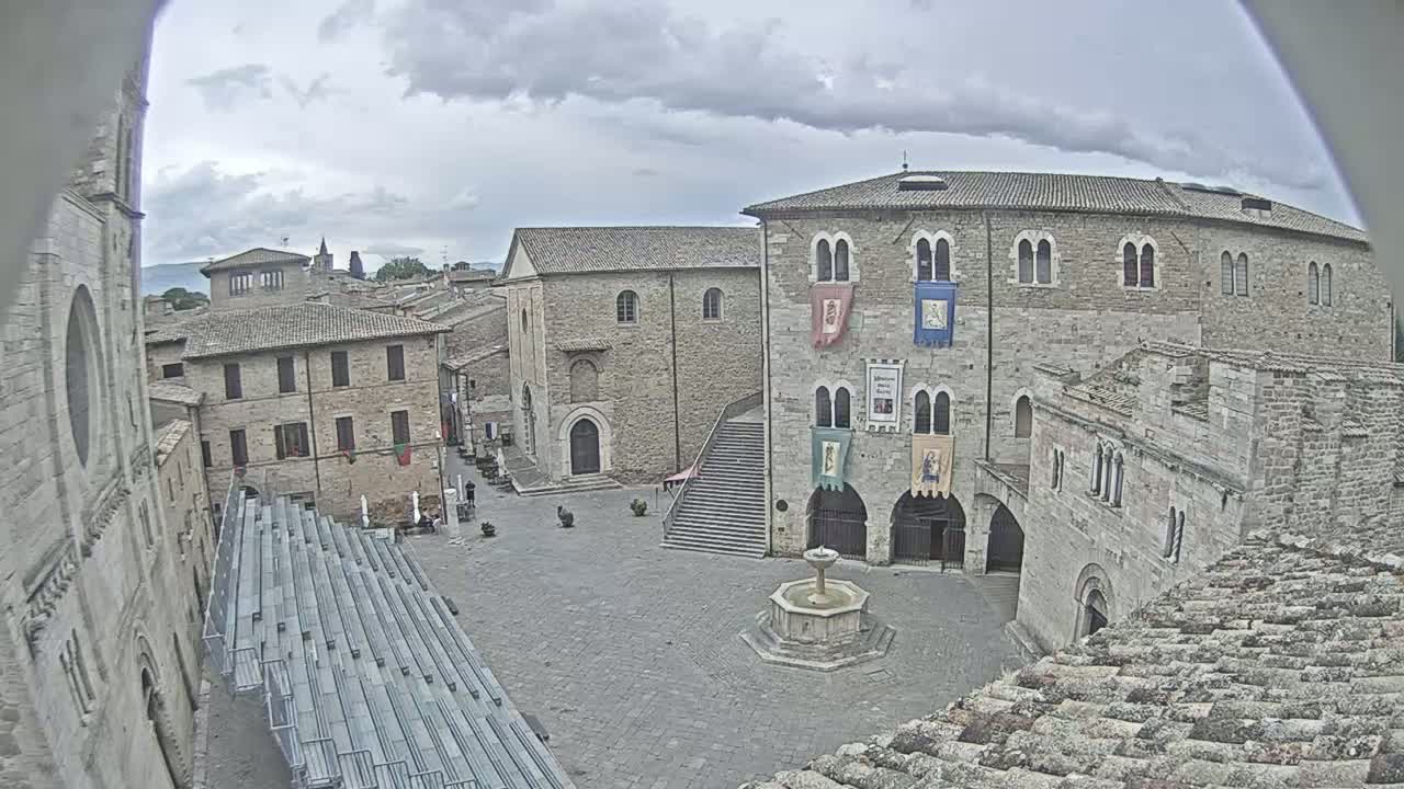 A town square in a European-style town under an overcast sky, featuring stone buildings, a fountain, and empty tiered seating.
