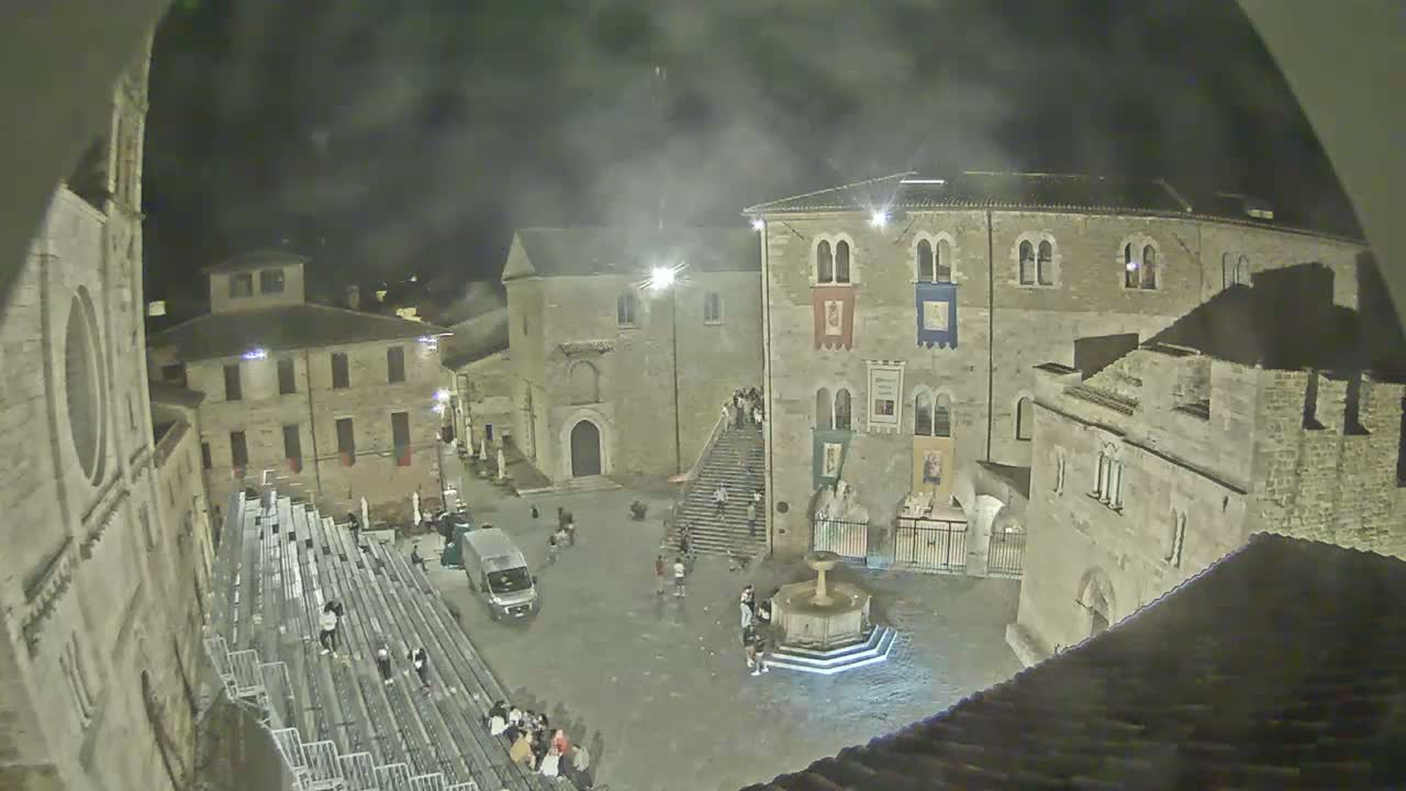 A nighttime view of a town square with several stone buildings, a fountain, bleachers, and a few people, under hazy or smoky conditions.