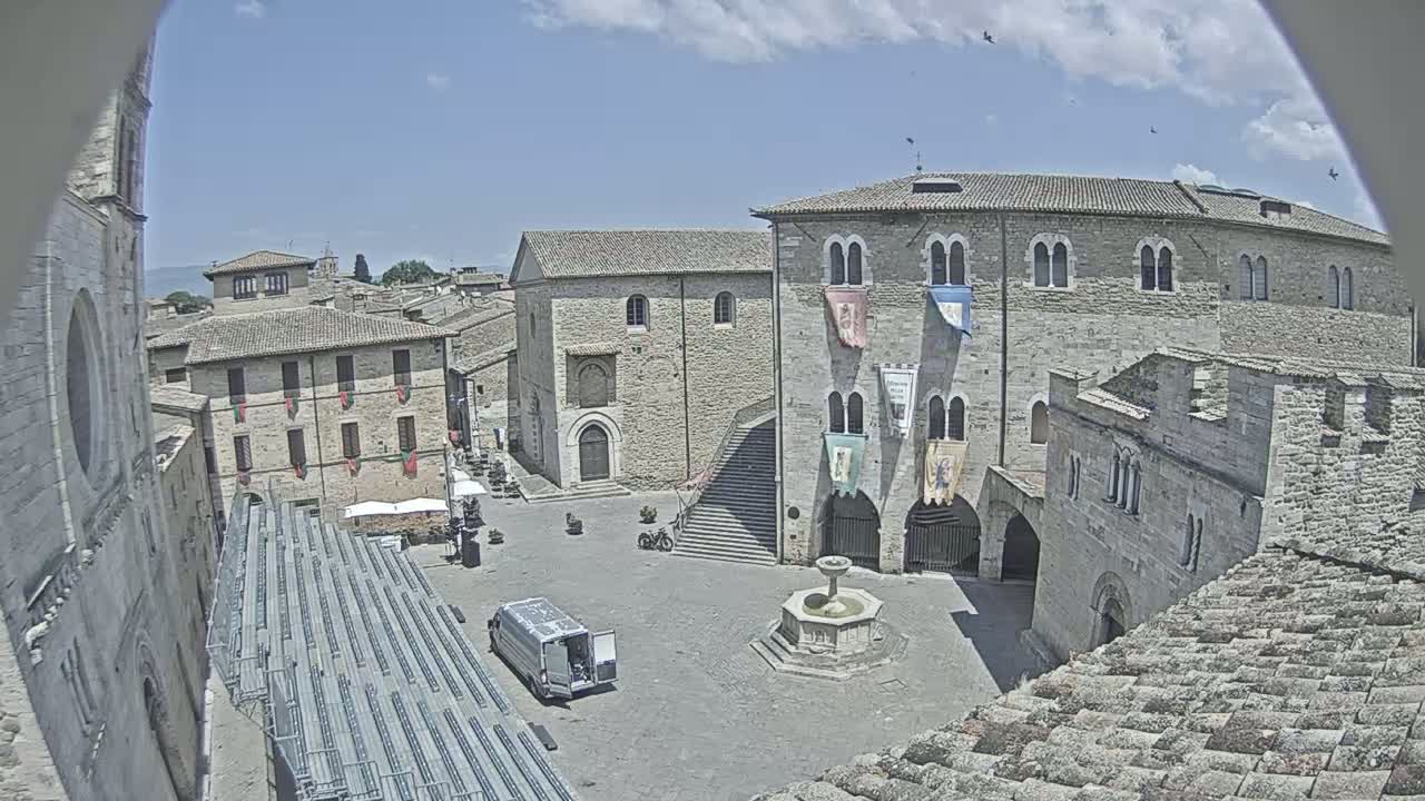 A mostly empty stone plaza with a fountain, surrounded by medieval-style buildings under a partly cloudy sky.
