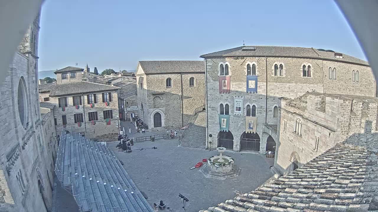 A high-angle view shows a town square in a historic European city on a sunny day, with stone buildings surrounding a paved plaza, featuring a fountain and temporary seating.