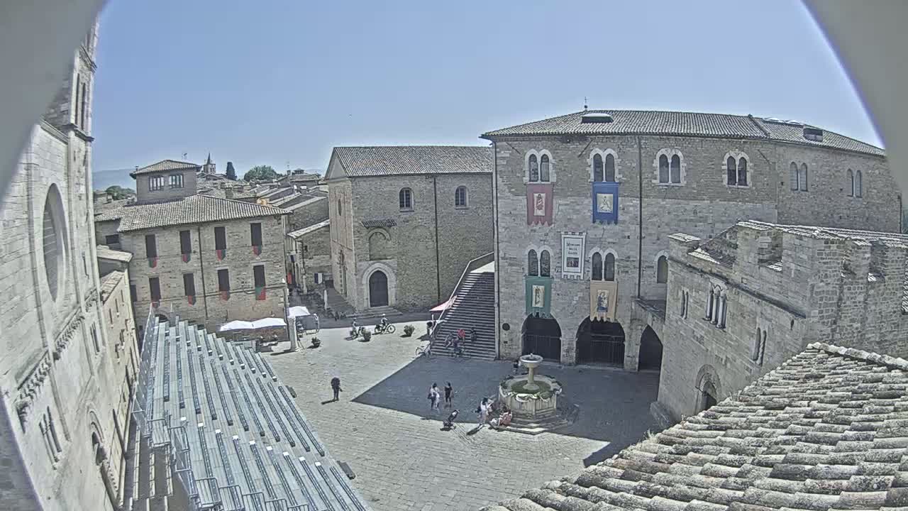 A sunny day reveals a town square in a historic European town, featuring stone buildings, a fountain, and a few people.