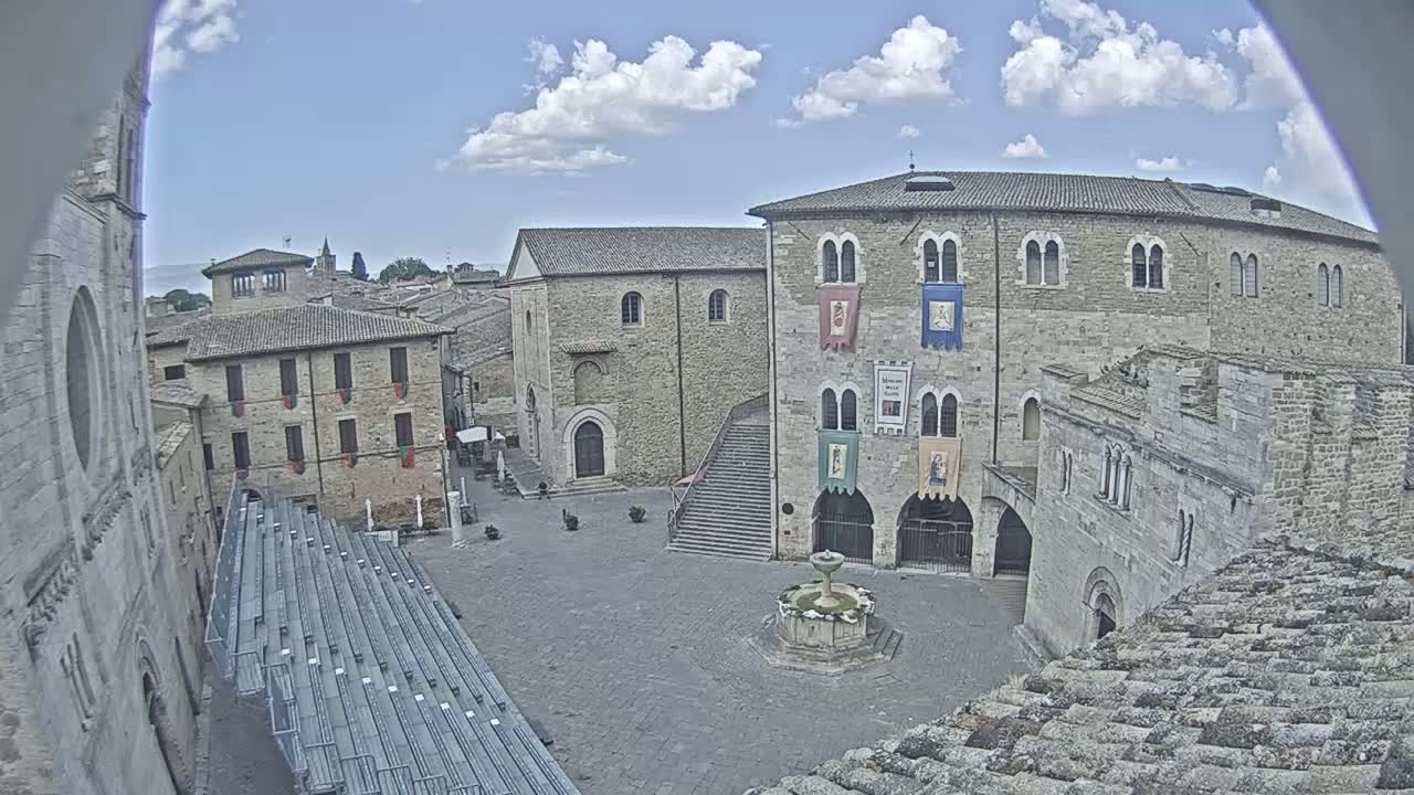 A partly cloudy sky overlooks a town square with medieval-style buildings, a fountain, and empty bleachers.