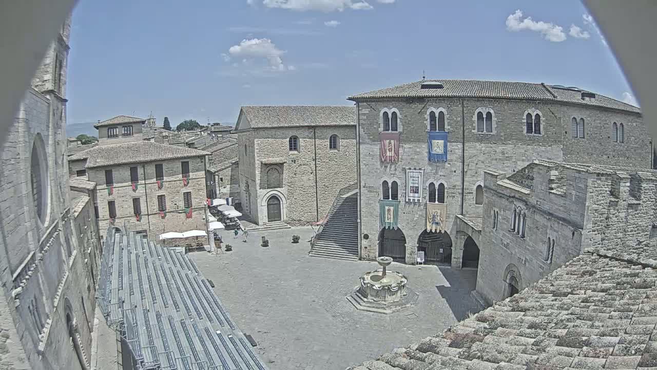 A sunny day reveals a town square in a historic European town, featuring stone buildings, a fountain, and empty seating.