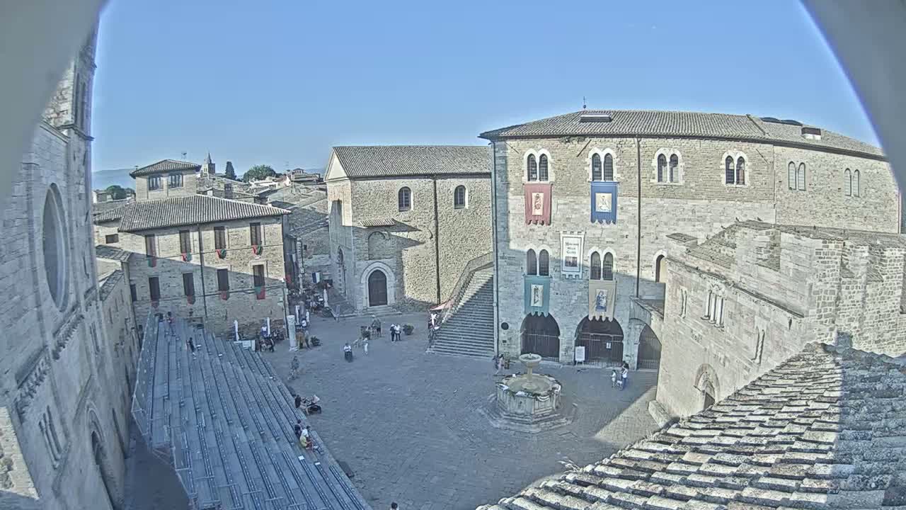 A sunny day reveals a town square surrounded by medieval-style buildings, including a large stone structure with banners, a fountain in the center, and tiered seating areas.
