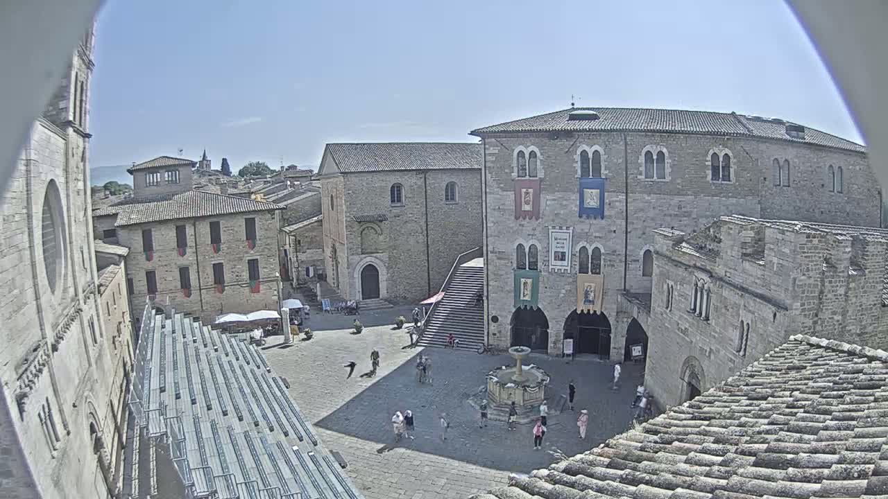 A sunny day reveals a town square with medieval-style buildings, a fountain, and a few people scattered throughout.