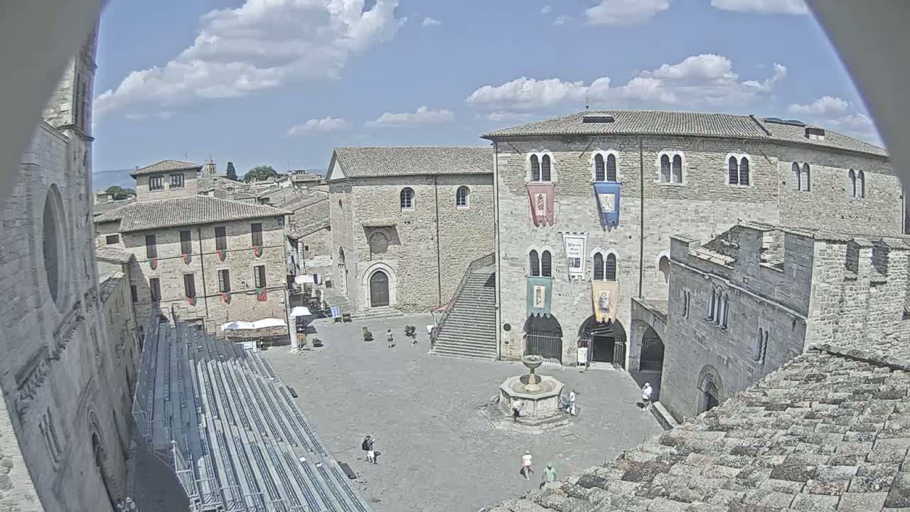 A mostly empty town square with stone buildings, a fountain, and bleachers under a partly cloudy sky.