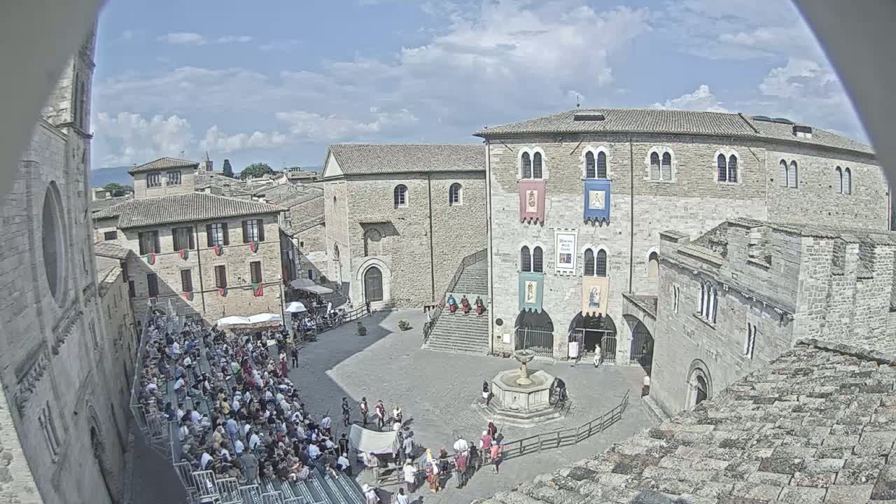 A large town square, under a partly cloudy sky, is filled with people sitting and standing around a fountain, surrounded by medieval-style buildings.