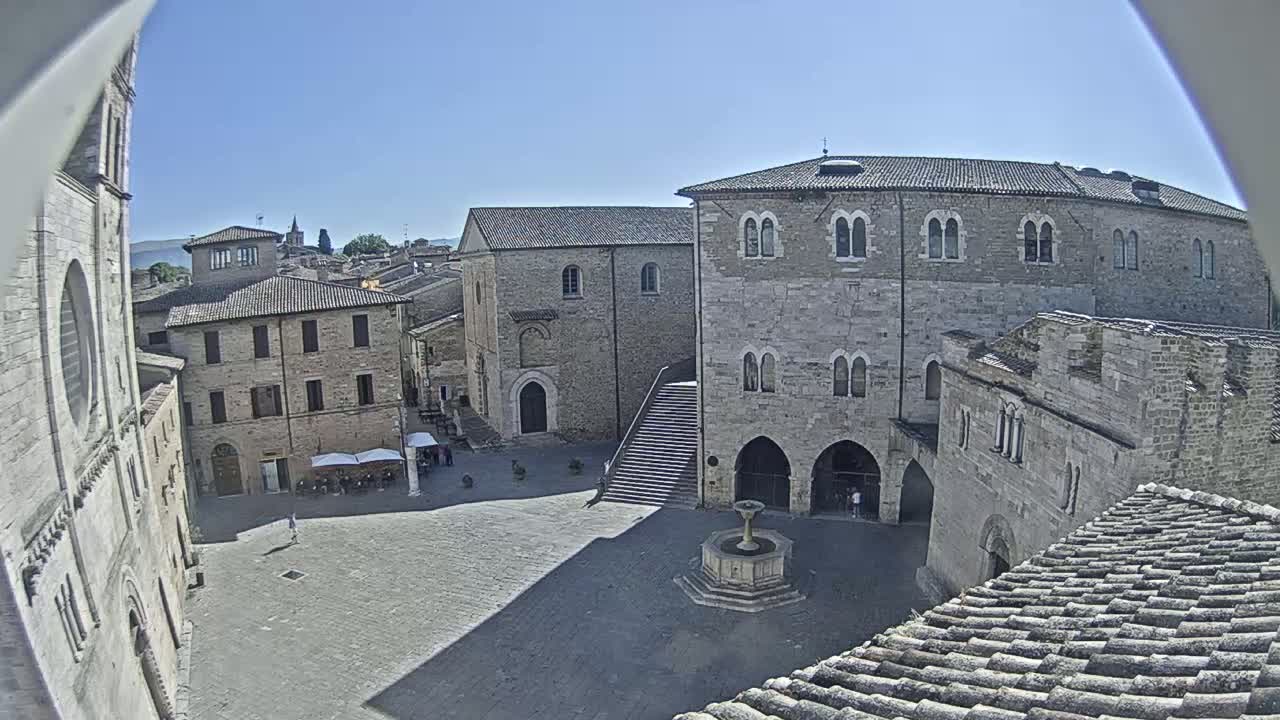 A stone plaza with a fountain, surrounded by medieval-style buildings under a clear blue sky.