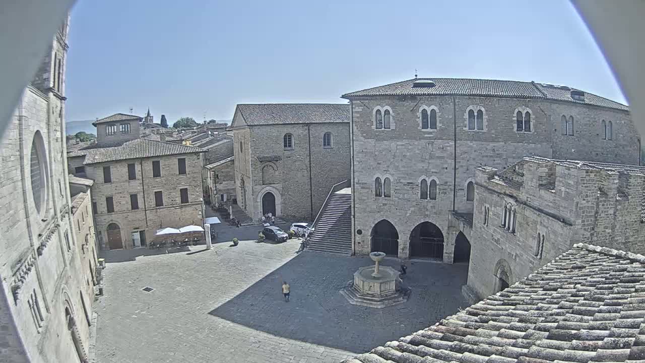 A sunny day reveals a town square surrounded by medieval stone buildings, including a large palazzo with a fountain in the center.
