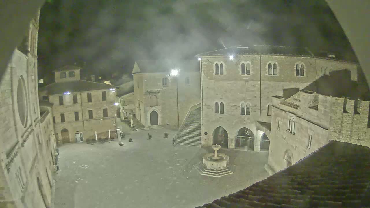 A sunny day reveals a town square surrounded by medieval stone buildings, including a large palazzo with a fountain in the center.