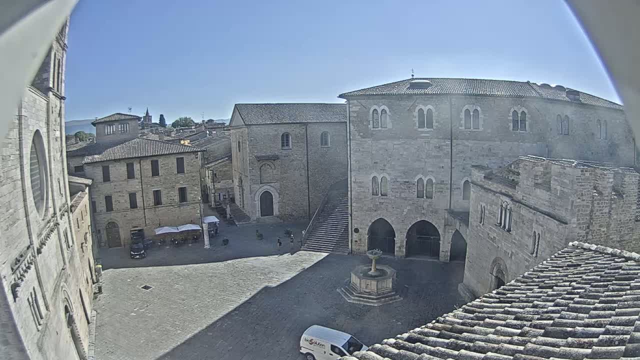 A high-angle view of a town square on a sunny day, surrounded by aged stone buildings, including a large palazzo and a church, with a fountain in the center.