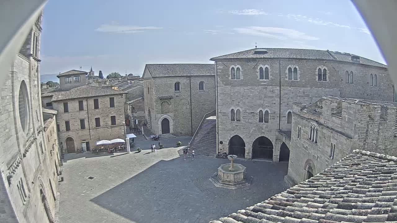 An elevated view shows a sunlit stone plaza with a fountain, surrounded by medieval-style buildings under a clear blue sky.