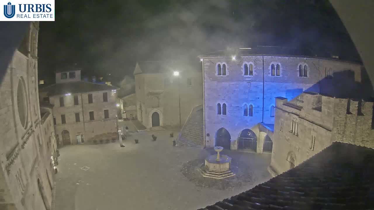 An empty European town square at night, featuring a central stone fountain and historic buildings, one dramatically lit in blue, appears wet and misty, suggesting light rain.