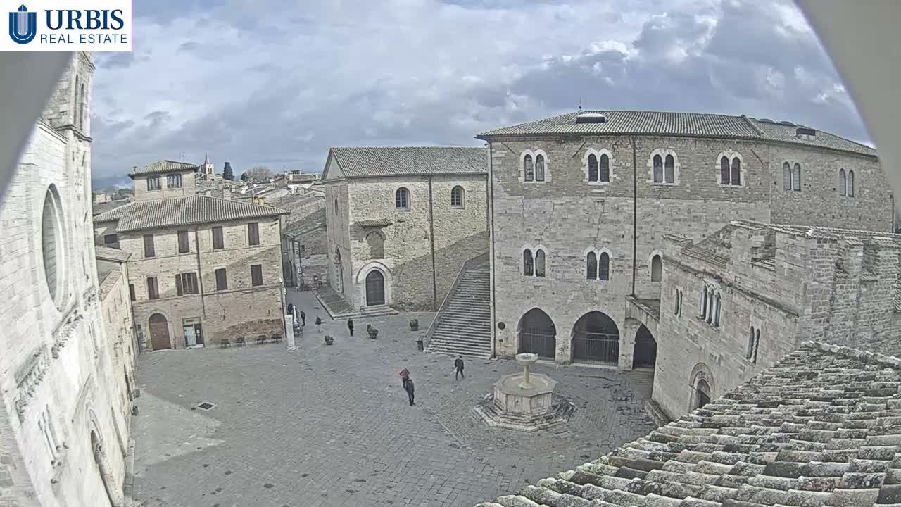 On a cloudy day, a wide-angle view captures a historic European town square with ancient stone buildings, a central tiered fountain, and several people walking across its cobblestone surface.