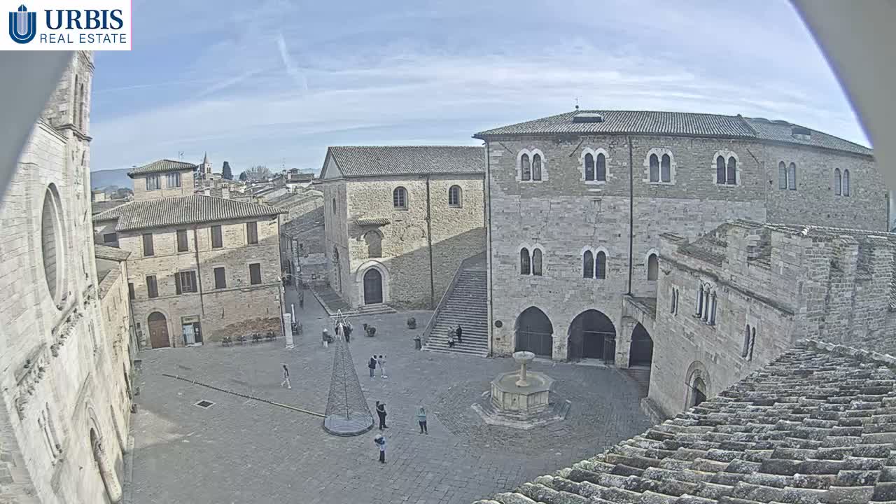 An elevated view captures a historic European town square featuring stone buildings, a central fountain, scattered pedestrians, and a conical modern art structure, under a partly cloudy sky.