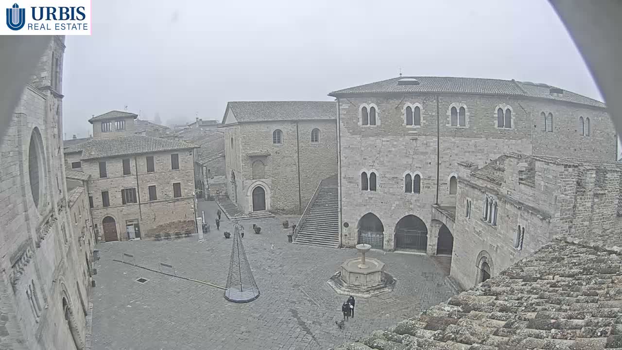 A quiet, old European stone town square is seen on a hazy, overcast day, featuring a central fountain, a tall conical structure, a grand stone staircase leading up to an ornate building, and numerous other historic stone buildings, with a few people scattered throughout.