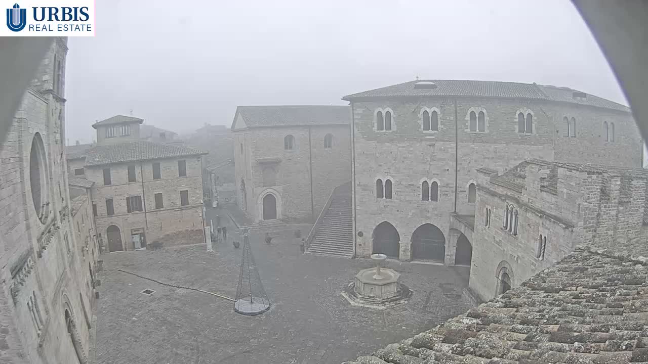 A quiet, old European stone town square is seen on a hazy, overcast day, featuring a central fountain, a tall conical structure, a grand stone staircase leading up to an ornate building, and numerous other historic stone buildings, with a few people scattered throughout.
