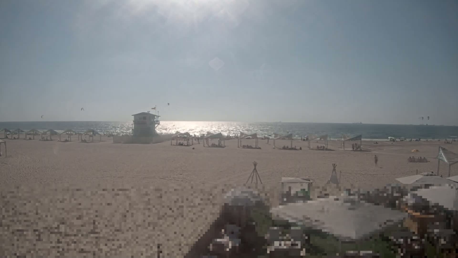 A sunny beach scene shows a lifeguard stand, numerous beach umbrellas, and people relaxing on a sandy beach next to a calm ocean with a few kitesurfers visible in the distance.
