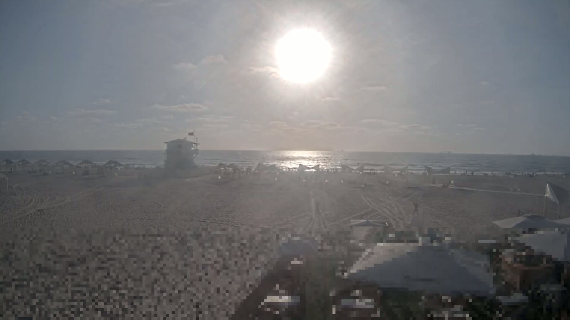 A sunny beach scene shows a lifeguard stand, numerous beach umbrellas, and people relaxing on a sandy beach next to a calm ocean with a few kitesurfers visible in the distance.