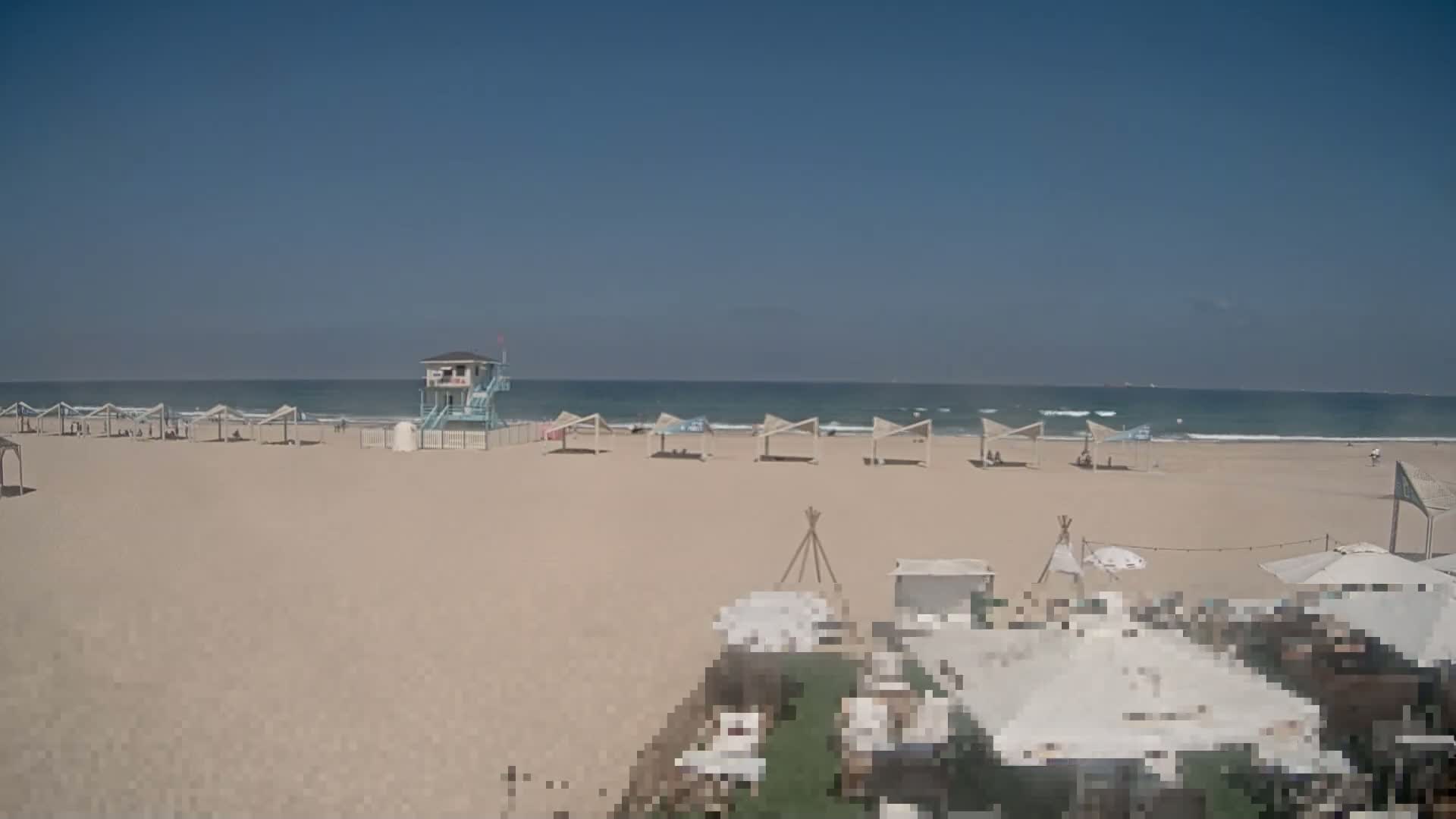 A sandy beach with several shaded structures and a lifeguard stand, under a clear blue sky and calm ocean waves.
