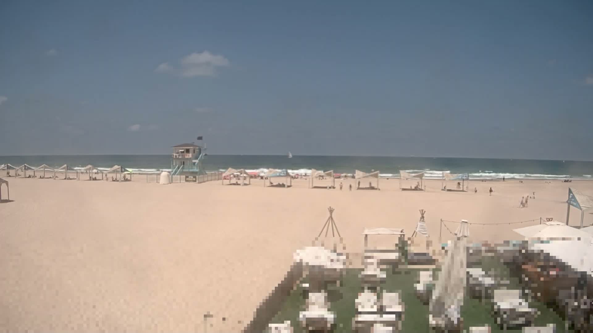 A sandy beach with scattered beachgoers and umbrellas under a mostly sunny sky, showing a lifeguard stand and ocean waves in the background.