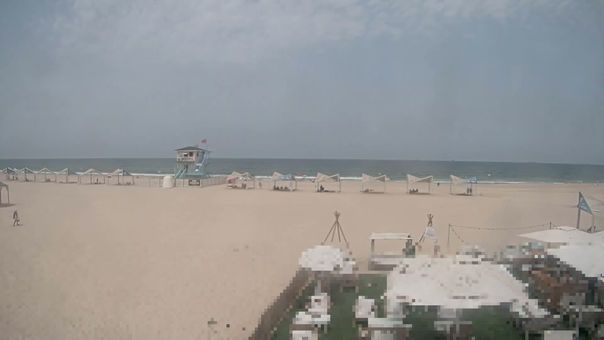 A sandy beach with several shaded structures, a lifeguard tower, and a calm ocean under an overcast sky is shown.
