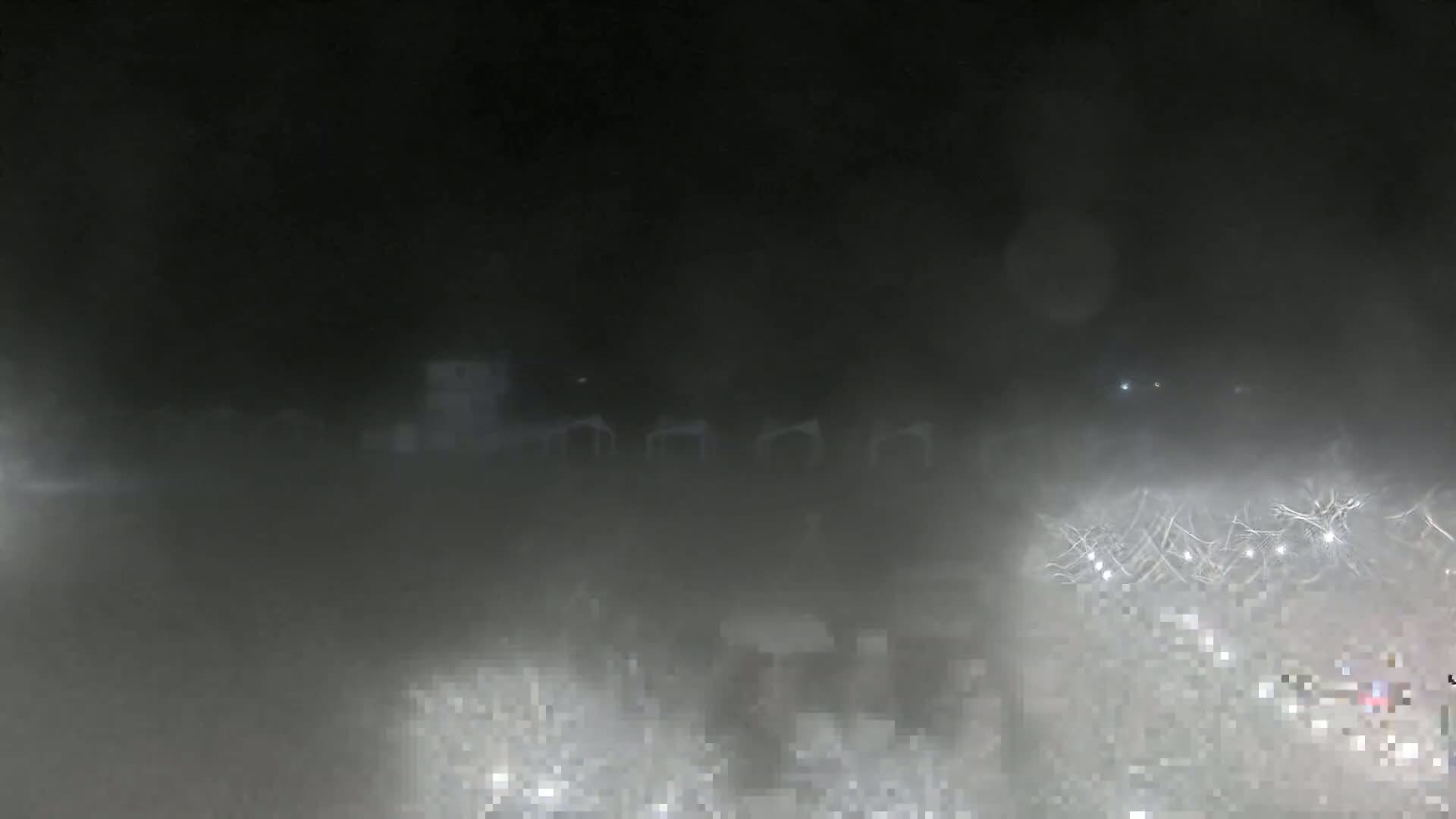 A nighttime scene is obscured by heavy fog, revealing only indistinct shapes of what appears to be structures and possibly vehicles in the distance.