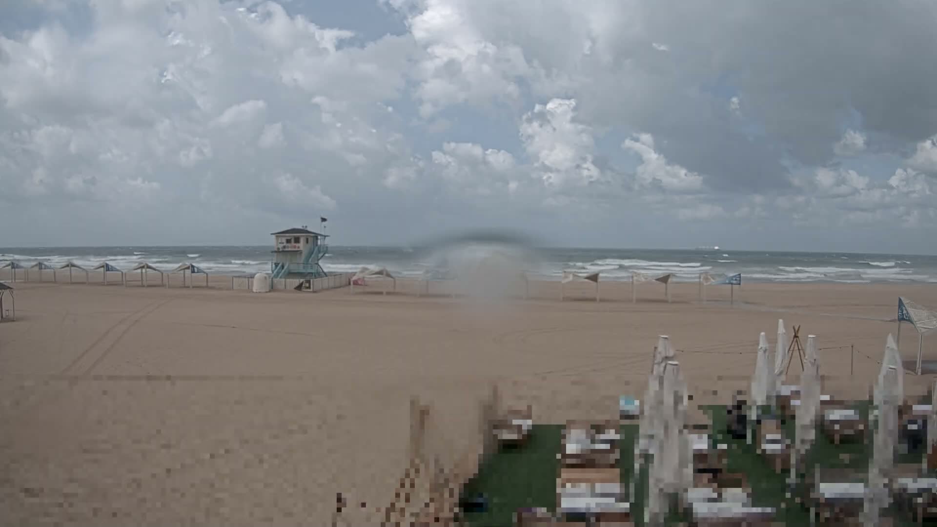 A wide sandy beach, featuring a lifeguard tower and many shelters, meets a wavy ocean under a partly cloudy sky, with a large, translucent blur dominating the center-midground of the view.