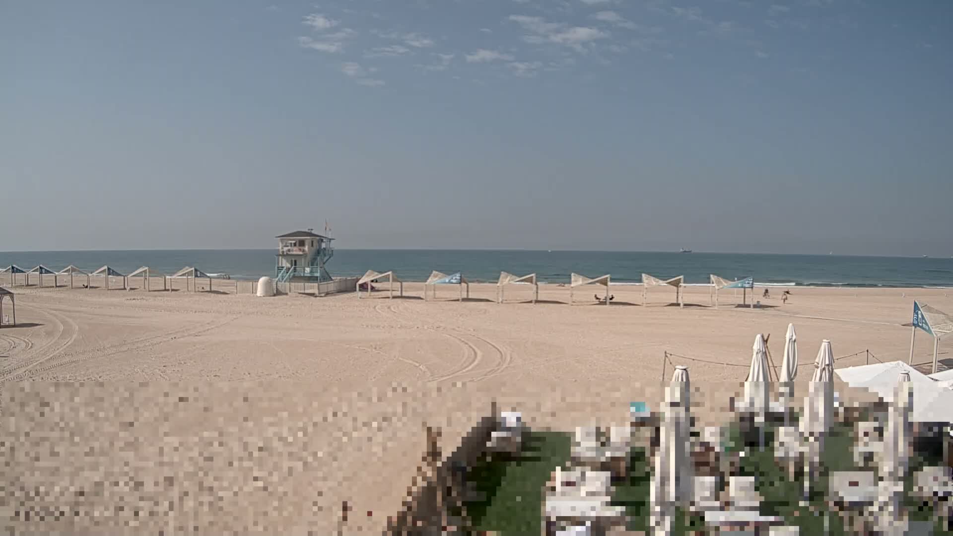 A wide sandy beach, featuring a lifeguard tower and many shelters, meets a wavy ocean under a partly cloudy sky, with a large, translucent blur dominating the center-midground of the view.