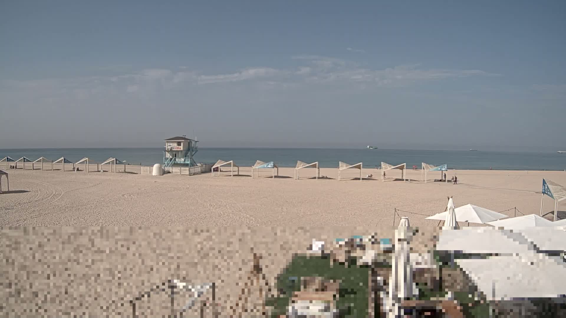 Under a clear and sunny sky, a wide sandy beach features a prominent blue and white lifeguard tower, a long row of geometric white sun shelters, and the calm blue ocean with distant boats, while a few people enjoy the shore.