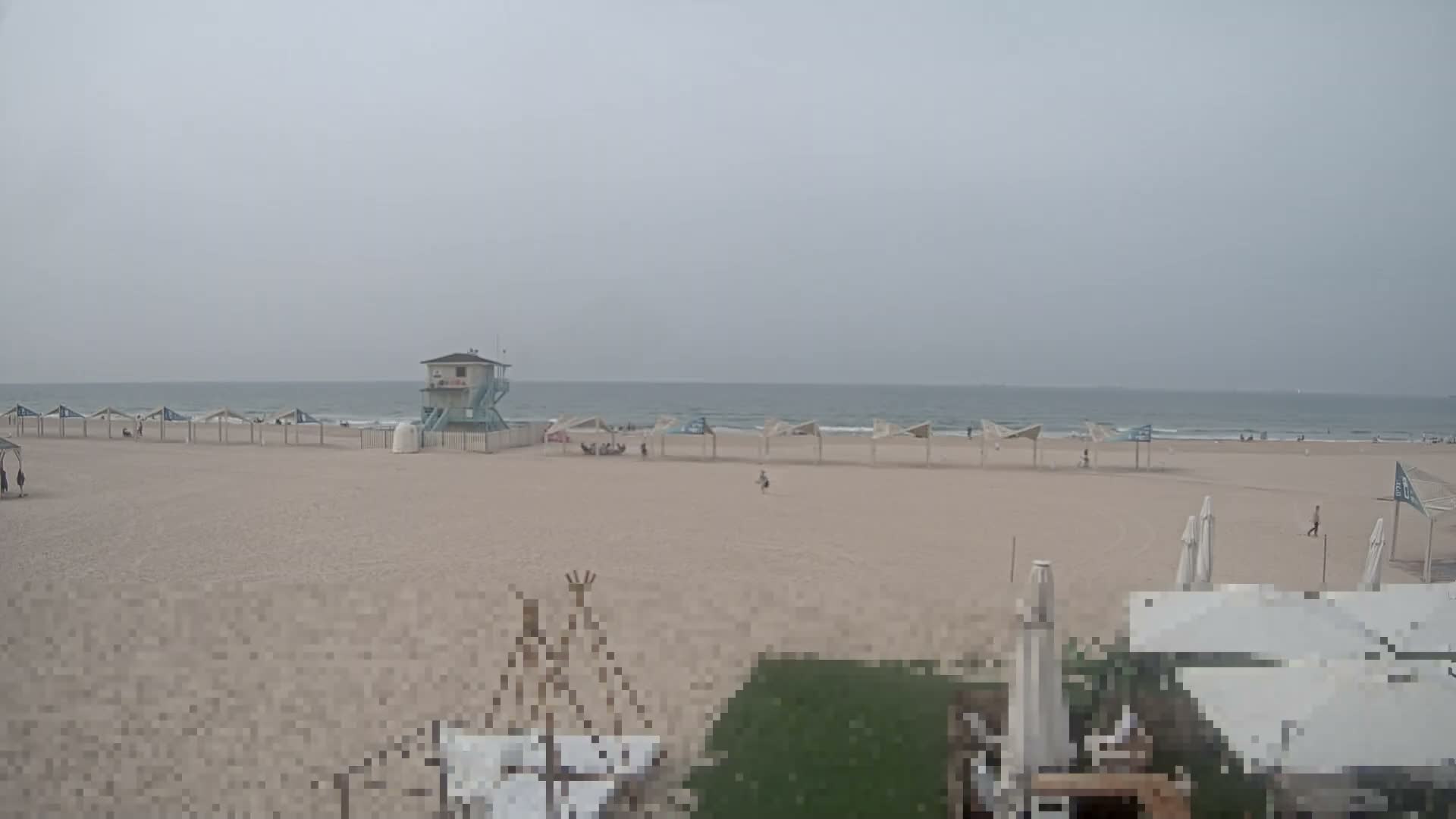 A wide sandy beach under an overcast sky features a light blue lifeguard tower, a long row of identical beach shelters stretching towards the ocean, and a few people scattered across the sand.