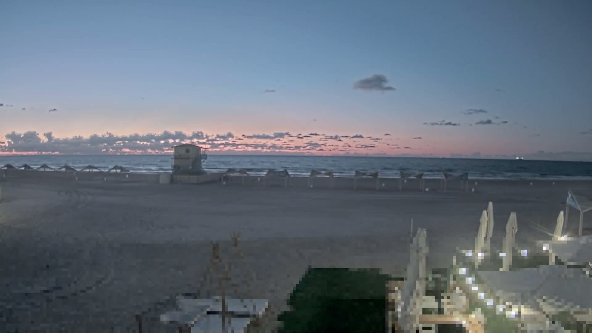 A tranquil beach scene unfolds at twilight under a clear, calm sky transitioning from blue to a warm pink horizon with scattered clouds, showcasing a sandy shore with a lifeguard tower and rows of beach umbrellas beside the ocean, while some foreground structures are illuminated.
