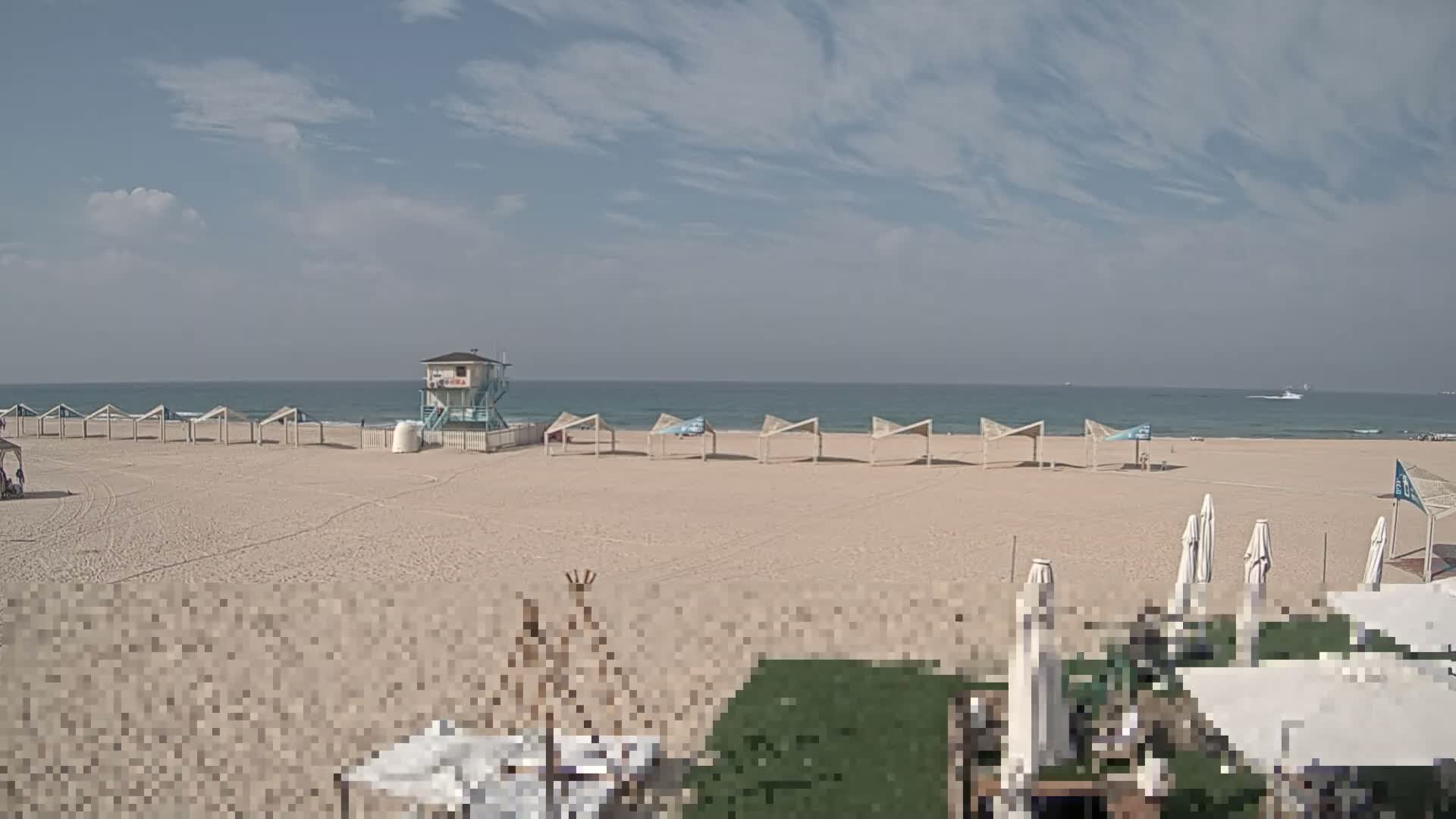 A wide, sandy beach extends to a calm blue ocean under a partly cloudy sky, featuring a prominent blue and white lifeguard tower, a line of light-colored open sun shelters, and several closed striped beach umbrellas in the foreground, with distant boats visible on the horizon.