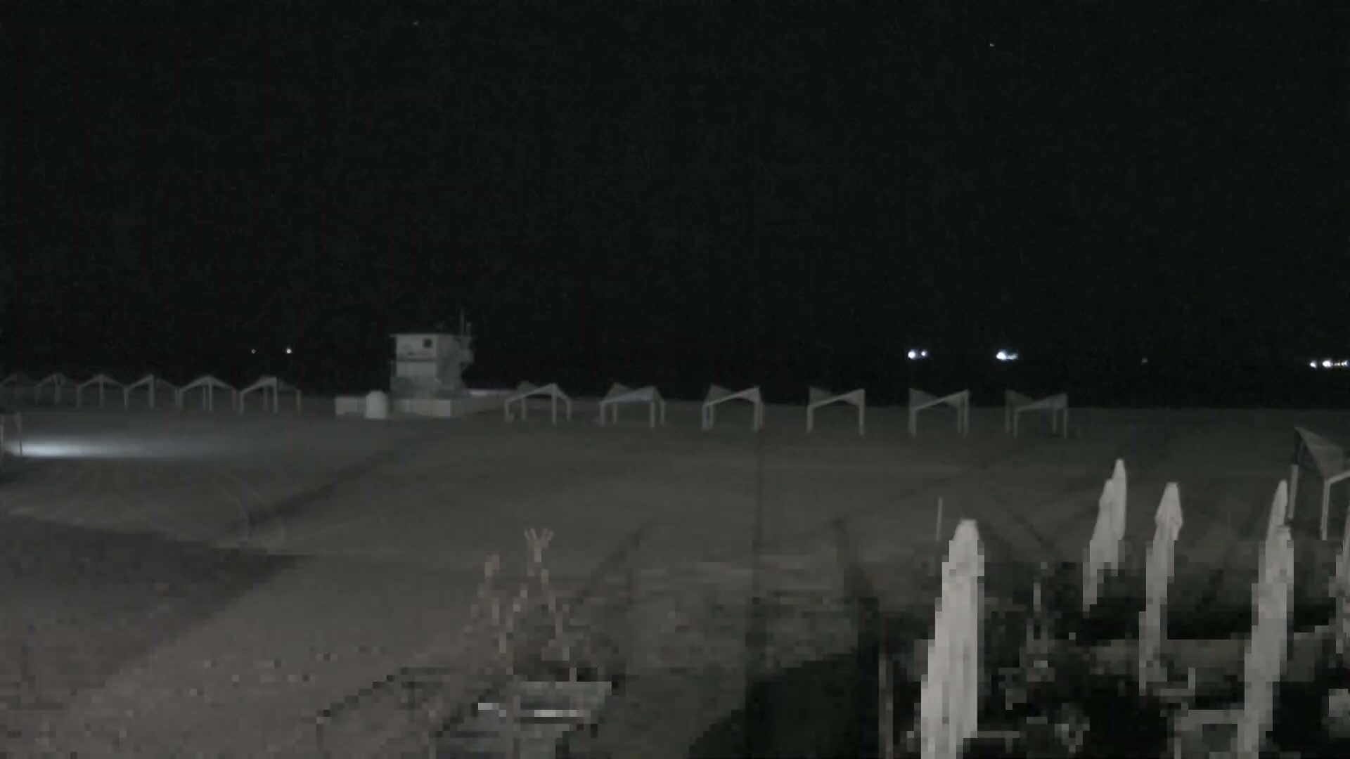 A wide, sandy beach extends to a calm blue ocean under a partly cloudy sky, featuring a prominent blue and white lifeguard tower, a line of light-colored open sun shelters, and several closed striped beach umbrellas in the foreground, with distant boats visible on the horizon.