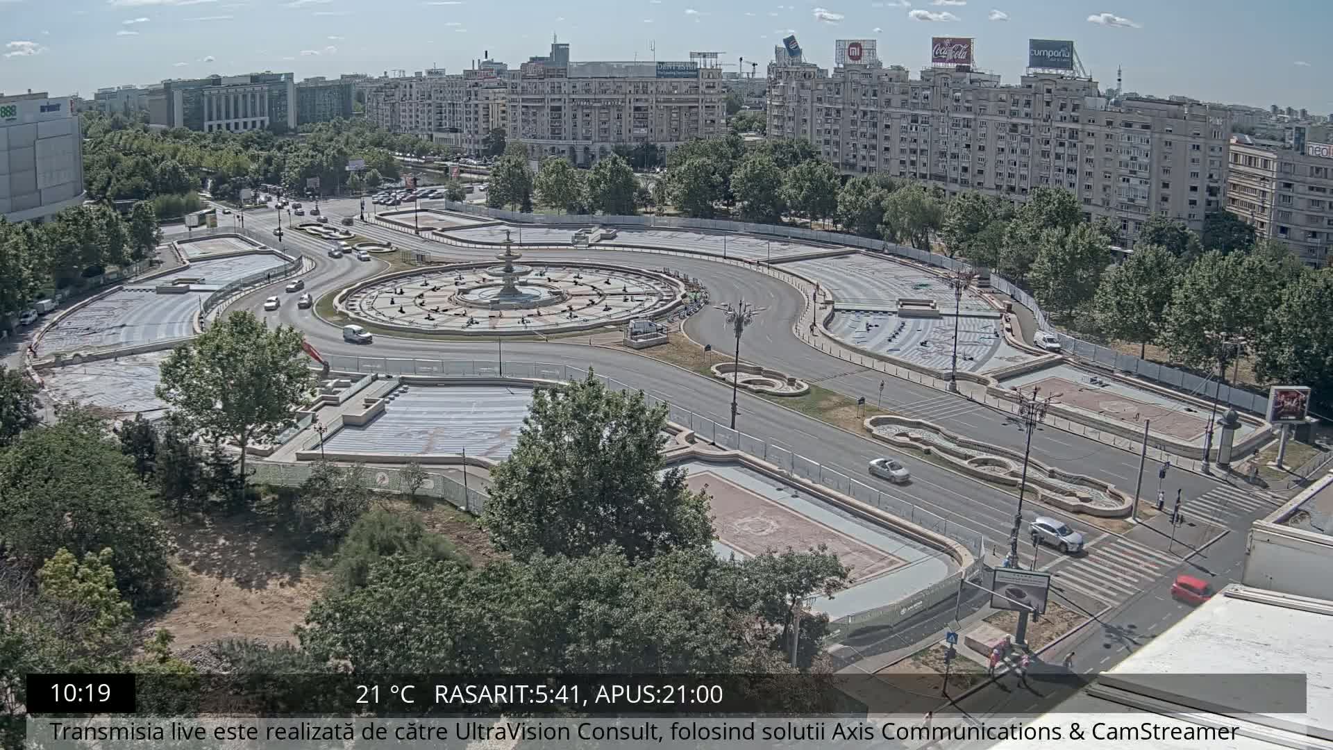 An aerial view of a city square under sunny conditions, showing a circular fountain surrounded by roadways and undergoing construction or renovation.