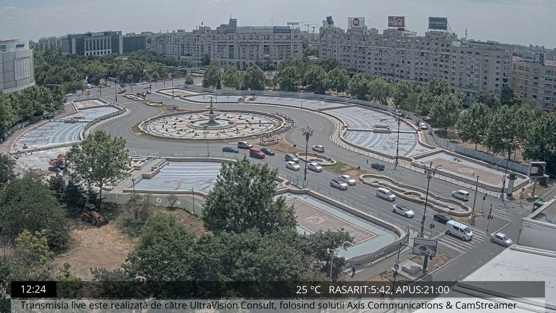 An aerial view of a city square under sunny conditions, showing a circular fountain surrounded by roadways and undergoing construction or renovation.