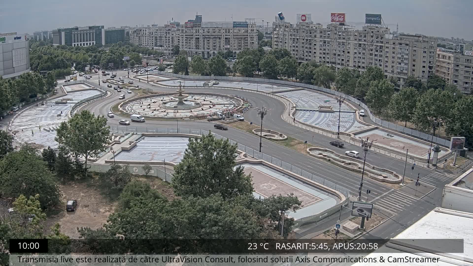 An aerial view shows a large, circular plaza under construction, with vehicles driving on surrounding roads and lush green trees in the surrounding area under a mostly clear sky.