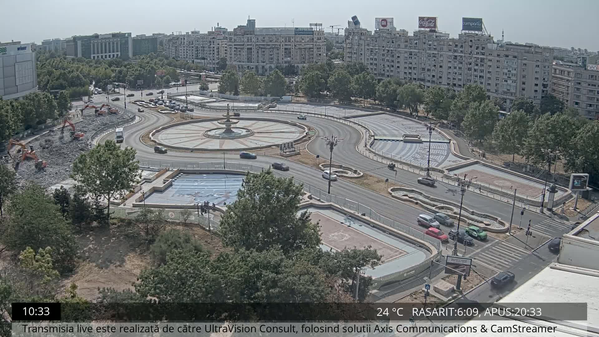 A high-angle, long shot on a sunny day shows a large roundabout under construction with several pieces of heavy machinery present and cars driving around the perimeter, surrounded by buildings and greenery.