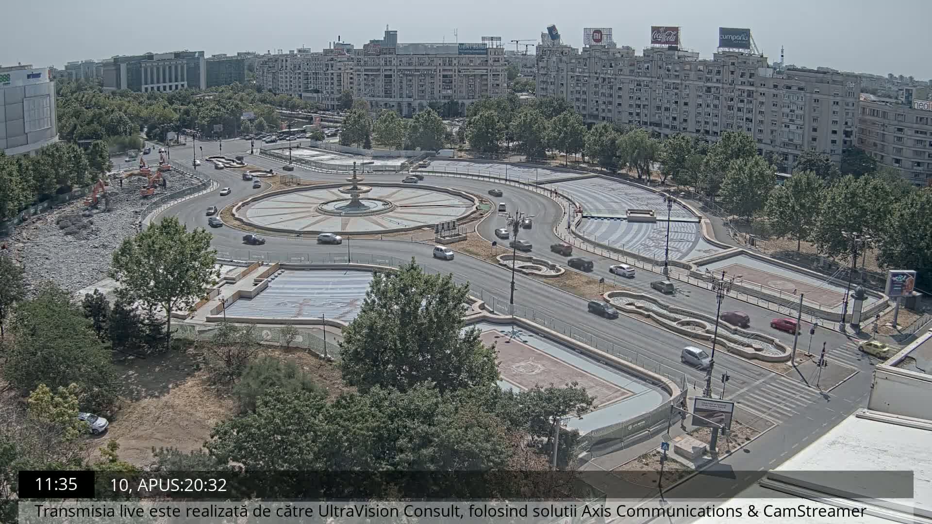 A high-angle view on a sunny day shows a city roundabout under construction, with vehicles moving around a central fountain and construction equipment visible in one section.