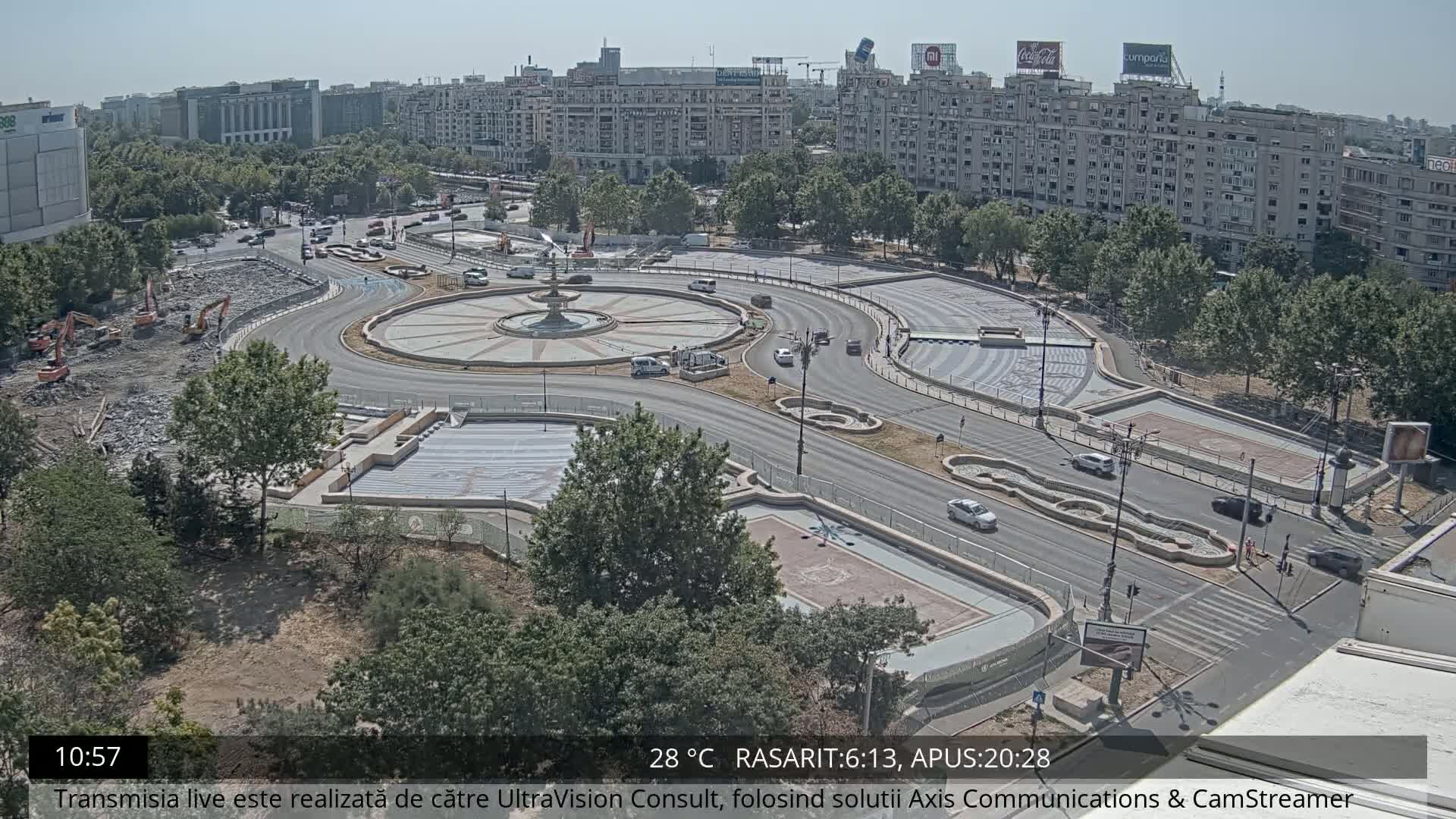A high-angle view shows a city roundabout under construction on a sunny day, with several vehicles driving around its perimeter and construction equipment visible in one section.