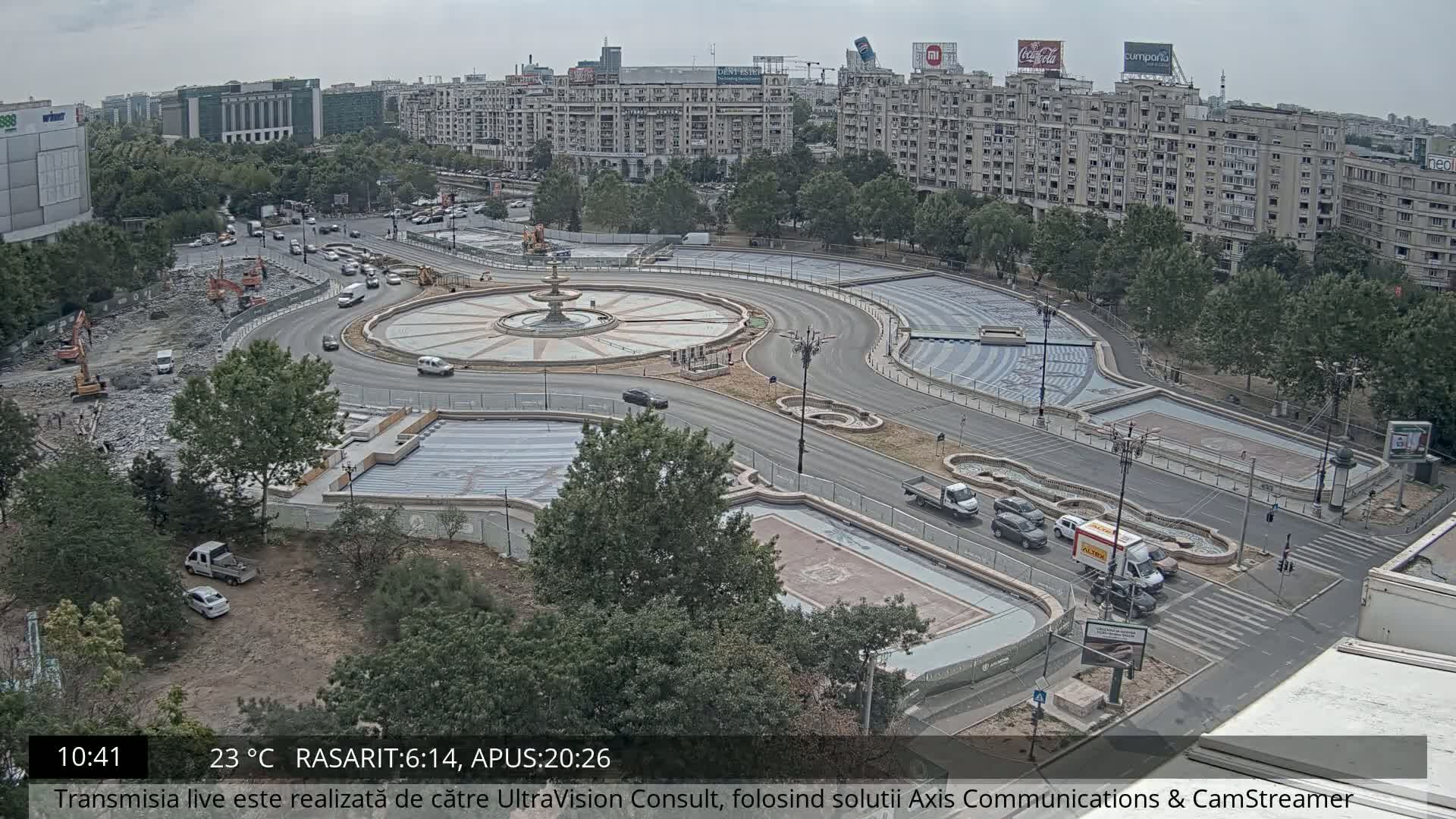 A high-angle view shows a city roundabout under construction on a partly cloudy day, with vehicles driving around the perimeter and construction equipment visible in the center.