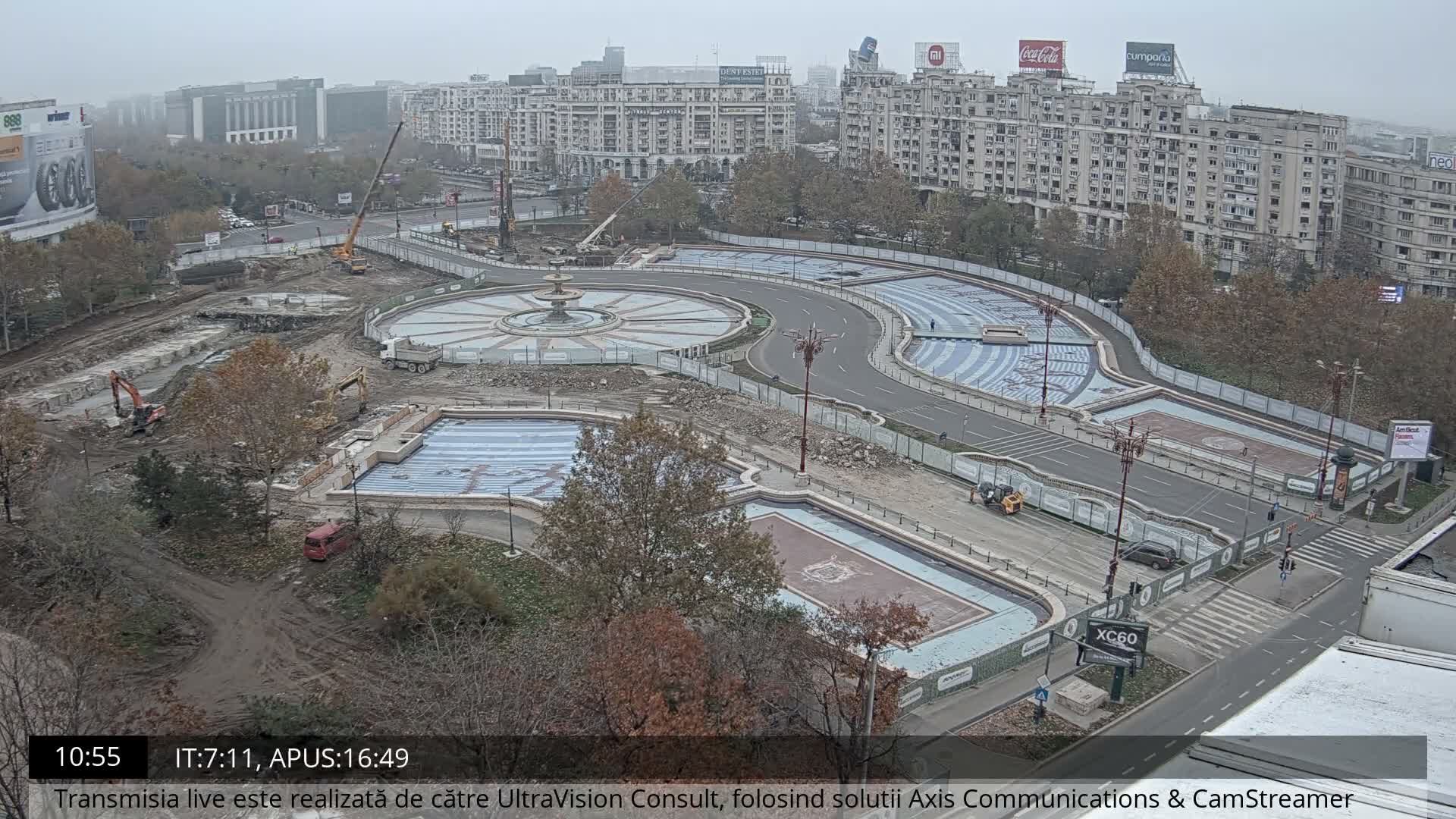 An aerial view reveals an extensive urban square undergoing renovation with multiple empty fountains and pools, active construction machinery, and surrounding city buildings, all under a hazy, overcast sky.