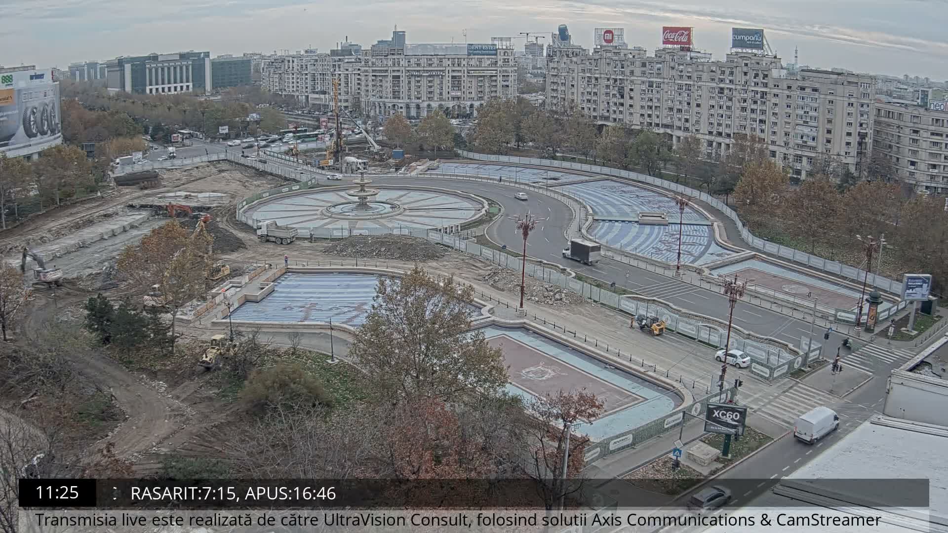 An aerial view reveals an extensive urban square undergoing renovation with multiple empty fountains and pools, active construction machinery, and surrounding city buildings, all under a hazy, overcast sky.
