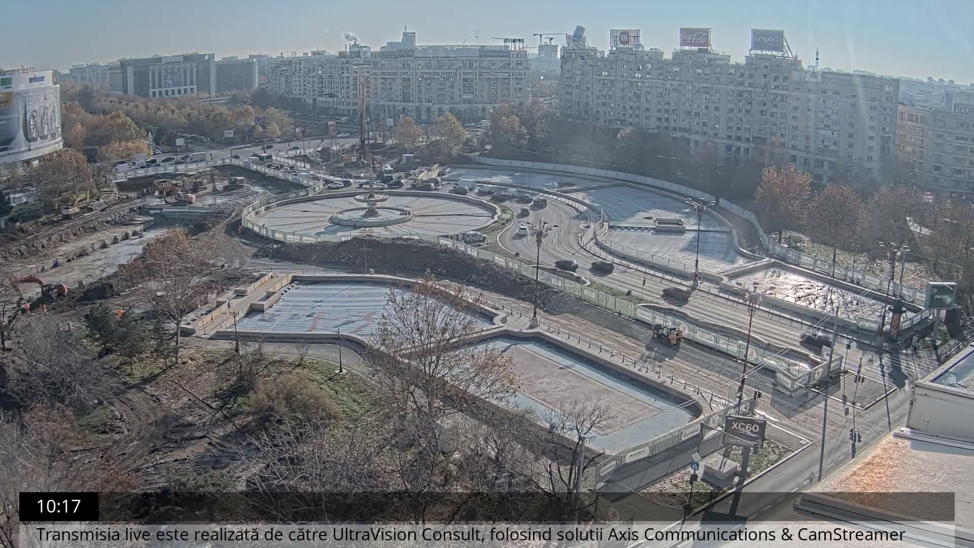 Under a hazy, cool sky, an aerial view captures a sprawling urban square undergoing extensive construction, featuring dirt mounds, excavators, empty fountain basins, and light vehicle traffic amidst numerous multi-story buildings in the background.