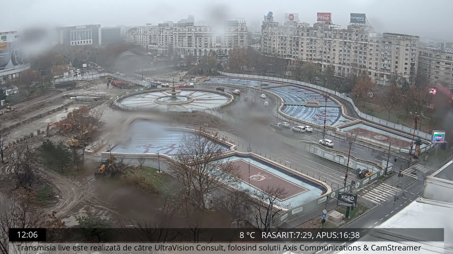 The image depicts a sprawling urban landscape under a gloomy, overcast, and damp sky, showcasing a large muddy construction site on the left, alongside a multi-tiered complex of ornate fountains and patterned basins, with roads carrying light traffic, all set against a backdrop of numerous city buildings.