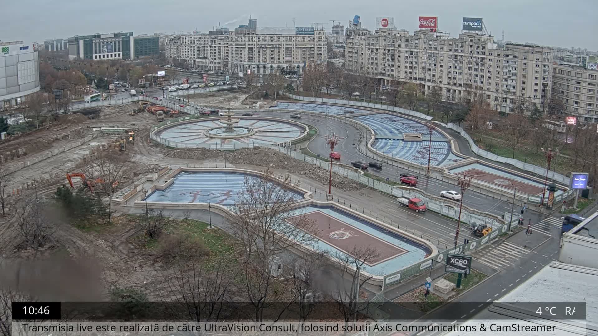 An aerial view captures a bustling city intersection undergoing construction, featuring multiple roads with moving traffic, several ornate but seemingly empty fountain pools, and numerous buildings under an overcast and damp sky.
