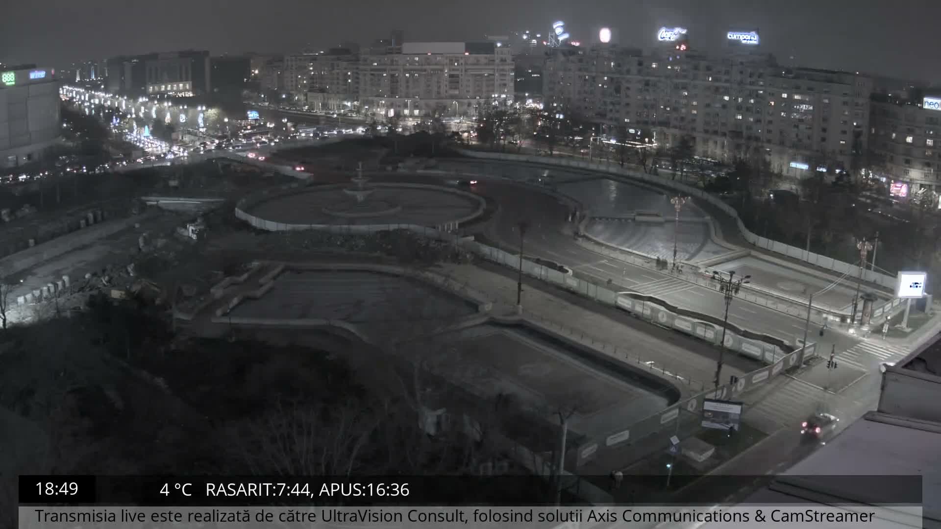 An aerial view captures a bustling city intersection undergoing construction, featuring multiple roads with moving traffic, several ornate but seemingly empty fountain pools, and numerous buildings under an overcast and damp sky.